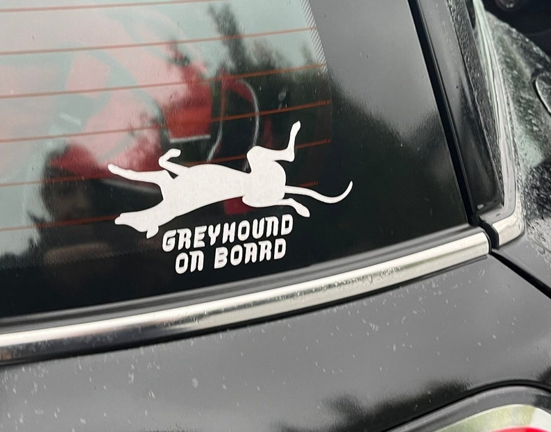 Peut inclure: Une silhouette blanche d'un l&eacute;vrier courant, avec le texte "Greyhound On Board" en dessous, sur une vitre de voiture noire.