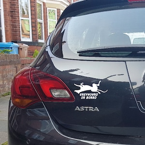 Peut inclure: Une voiture noire avec un autocollant blanc qui dit "Greyhound on Board" et une silhouette blanche d'un l&eacute;vrier courant. La voiture est une Opel Astra.