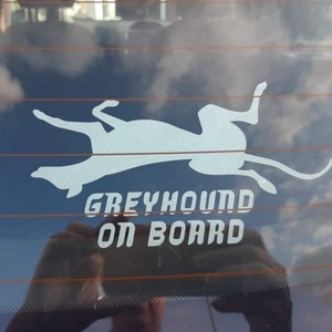 Peut inclure: Une silhouette blanche d'un l&eacute;vrier couch&eacute; sur le dos sur la vitre arri&egrave;re d'une voiture bleue. Le texte "GREYHOUND ON BOARD" est sous le chien.