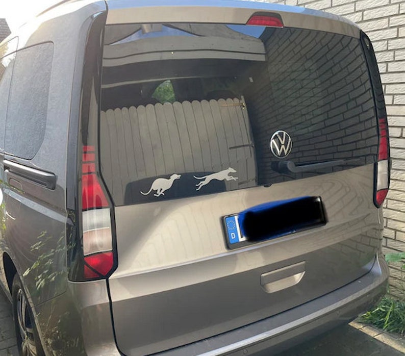 Puede incluir: Una minivan gris con una ventana trasera negra y un logotipo de Volkswagen. La ventana trasera tiene dos siluetas de perros grises corriendo en el vidrio. El coche tiene una matr&iacute;cula con la letra D.