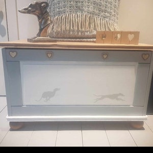 Puede incluir: Una caja de almacenamiento de madera azul claro con un interior blanco y detalles decorativos en forma de coraz&oacute;n. La caja tiene un borde blanco y se apoya en cuatro patas de madera. Dos siluetas de galgos est&aacute;n pintadas en la parte delantera de la caja.