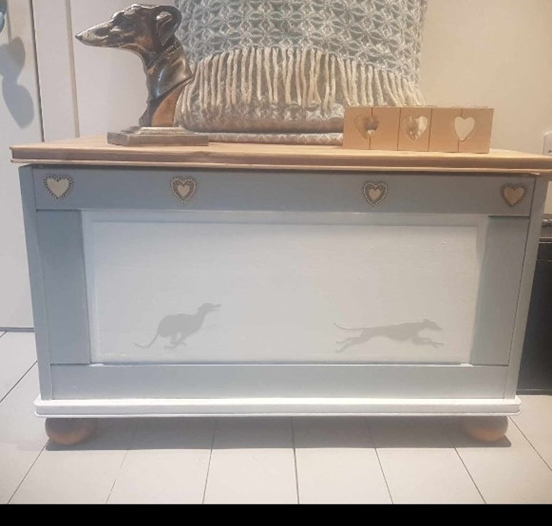 Puede incluir: Una caja de almacenamiento de madera azul claro con un interior blanco y detalles decorativos en forma de coraz&oacute;n. La caja tiene un borde blanco y se apoya en cuatro patas de madera. Dos siluetas de galgos est&aacute;n pintadas en la parte delantera de la caja.