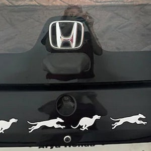 Puede incluir: Un coche negro Honda Brio i-VTEC con un logotipo Honda plateado en la parte trasera. El coche tiene cuatro pegatinas de silueta blanca de perros corriendo en el parachoques trasero.