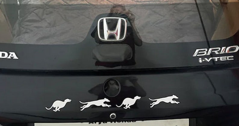 Puede incluir: Un coche negro Honda Brio i-VTEC con un logotipo Honda plateado en la parte trasera. El coche tiene cuatro pegatinas de silueta blanca de perros corriendo en el parachoques trasero.