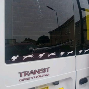 Puede incluir: Una furgoneta blanca con las palabras "TRANSIT GREYHOUND" en la parte trasera. La furgoneta tiene una ventana negra con el reflejo de un edificio y una farola. Hay varias siluetas blancas de galgos corriendo a trav&eacute;s de la ventana trasera.
