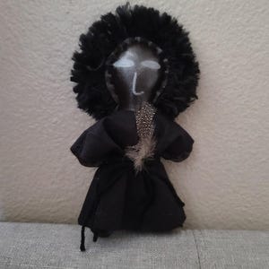 May include: A handcrafted black doll featuring a feathery head covering and a black dress. The doll's face is painted with white eyes and a simple nose. A single feather is attached to the dress.