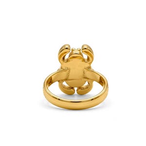 May include: A gold-toned ring featuring a stylised crab design on the face. The band is smooth and circular, while the crab motif displays rounded pincers and legs extending from the central body. The entire piece has a polished, reflective finish.