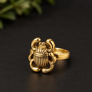 May include: A gold-toned ring featuring a detailed scarab beetle design as the centrepiece. The beetle has a smooth, rounded head and a textured, ribbed body with curved legs extending outward. The band of the ring is plain and polished.