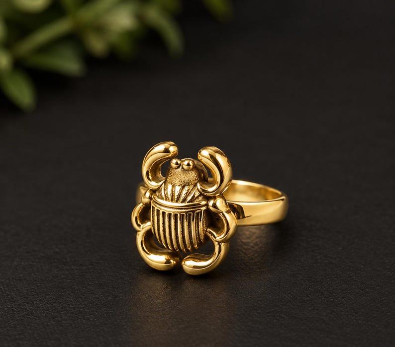 May include: A gold-toned ring featuring a detailed scarab beetle design as the centrepiece. The beetle has a smooth, rounded head and a textured, ribbed body with curved legs extending outward. The band of the ring is plain and polished.
