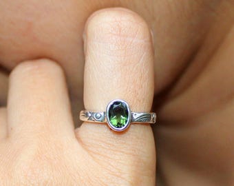 May include: A silver ring featuring an oval, green gemstone. The ring has a detailed band with leaf and dot patterns. The gemstone is set in a silver bezel. The ring is shown on a finger.