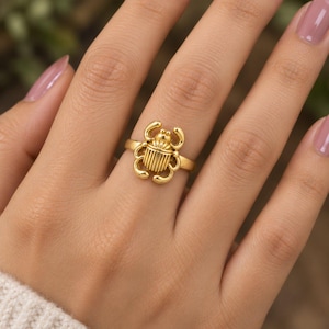 May include: A gold-toned ring featuring a central scarab beetle motif with a smooth, polished finish. The beetle design includes detailed vertical lines on the shell and curved legs extending from the sides, set on a simple, thin, circular band.