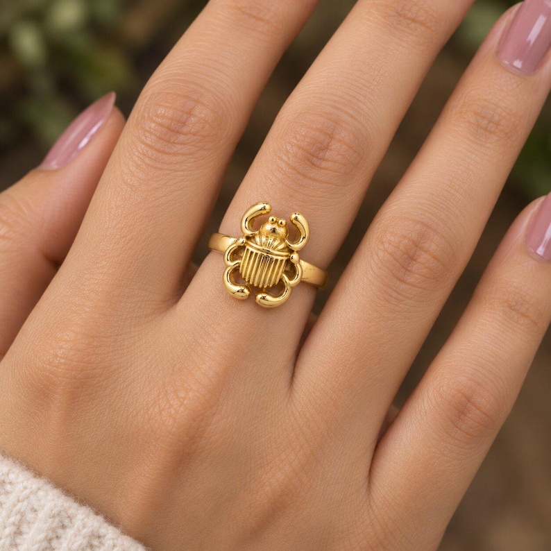 May include: A gold-toned ring featuring a central scarab beetle motif with a smooth, polished finish. The beetle design includes detailed vertical lines on the shell and curved legs extending from the sides, set on a simple, thin, circular band.