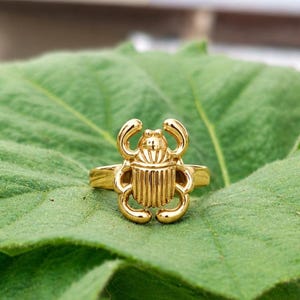 May include: A gold-toned ring featuring a detailed scarab beetle design as the centrepiece. The beetle has a smooth, polished surface with vertical ridges on its back and curved legs extending from the sides, attached to a simple, plain metal band.