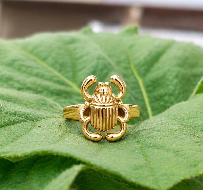 May include: A gold-toned ring featuring a detailed scarab beetle design as the centrepiece. The beetle has a smooth, polished surface with vertical ridges on its back and curved legs extending from the sides, attached to a simple, plain metal band.