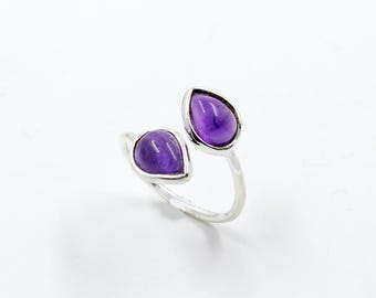 May include: A silver ring featuring two teardrop-shaped, vibrant purple gemstones. The gemstones are set in a polished silver band, creating an open design. The ring is displayed against a white background, highlighting its elegant simplicity.