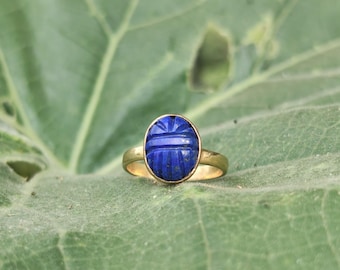 May include: A gold ring featuring an oval-shaped, deep blue lapis lazuli gemstone carved with horizontal lines. The stone is set in a simple gold band, creating a contrast between the vibrant blue of the gem and the metallic gold setting.