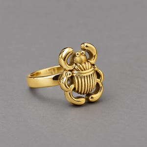 May include: A gold-toned ring featuring a three-dimensional scarab beetle design. The beetle has a polished, reflective surface with detailed, vertical ridges on its back and curved legs extending from the sides of the central body element.