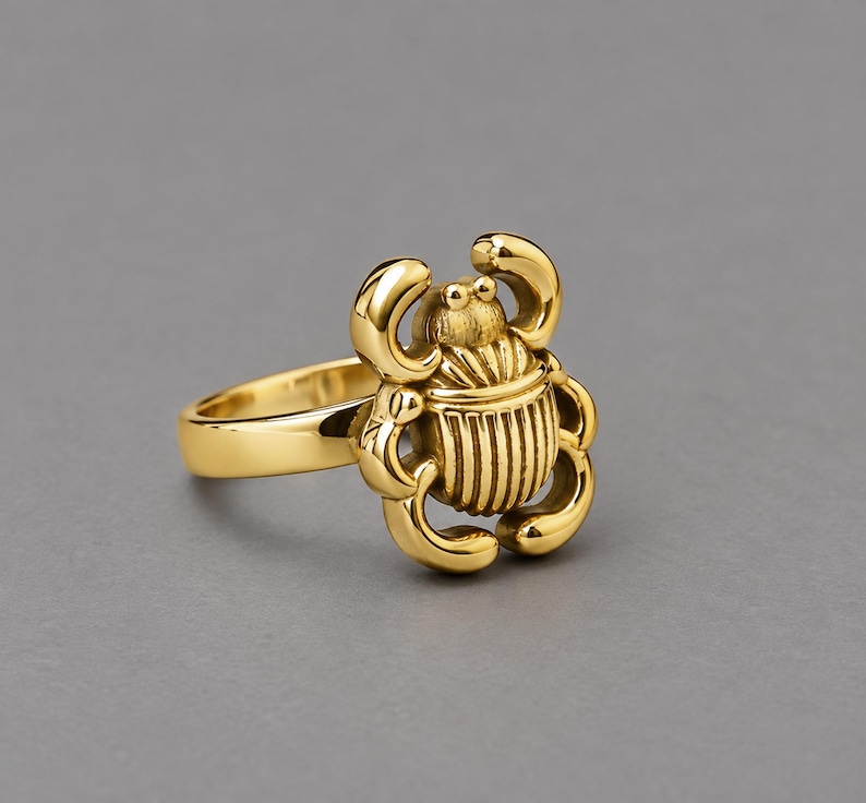 May include: A gold-toned ring featuring a three-dimensional scarab beetle design. The beetle has a polished, reflective surface with detailed, vertical ridges on its back and curved legs extending from the sides of the central body element.