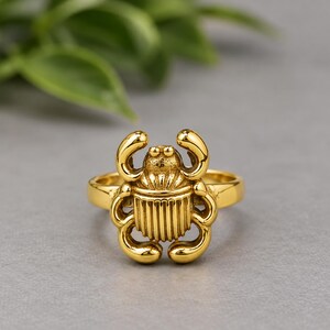 May include: A gold-toned ring featuring a central scarab beetle motif. The beetle design includes detailed vertical lines on the body, curved legs, and a textured head section. The band of the ring is smooth and circular, maintaining a consistent gold finish throughout the entire piece of jewellery.