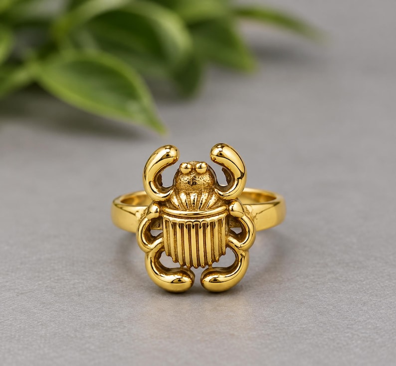 May include: A gold-toned ring featuring a central scarab beetle motif. The beetle design includes detailed vertical lines on the body, curved legs, and a textured head section. The band of the ring is smooth and circular, maintaining a consistent gold finish throughout the entire piece of jewellery.