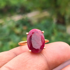 May include: A gold ring featuring a large, oval-cut, deep red gemstone set in a four-prong mounting. The faceted stone displays a rich crimson hue with natural inclusions visible across its surface, held securely by a thin, polished gold band.