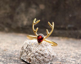 May include: A gold-toned ring featuring a metal deer antler design and a single, pear-shaped, deep red gemstone set at the base of the antlers. The ring band is thin and circular, showcasing a polished metallic finish throughout the structure.