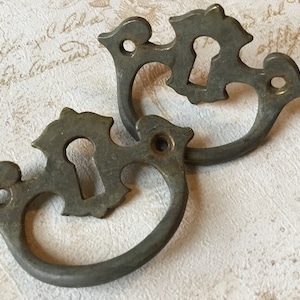 May include: Two antique bronze drawer pulls. Each pull has a decorative top with a keyhole and a curved handle. The pulls have a vintage, aged appearance, suggesting they are reclaimed or vintage hardware.