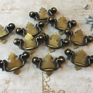 May include: Ten brass-effect drawer pulls with a decorative design. Each pull has a curved handle and ornate backplate. Small silver screws are scattered nearby, suggesting a hardware set for furniture restoration or new construction.