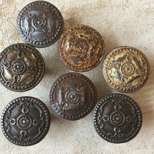 May include: A collection of seven antique metal knobs with a circular design. The knobs feature a central floral pattern surrounded by ornate detailing and are finished in various shades of bronze, gold, and silver.