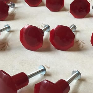 May include: A collection of red octagonal cabinet knobs with silver screws. The knobs are made of a shiny, glossy material.