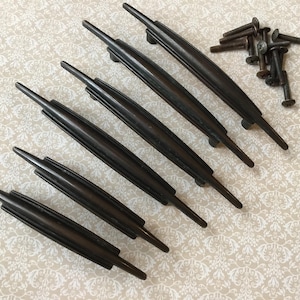 May include: A set of six dark brown cabinet pulls with a ribbed center design, along with a pile of matching screws. The pulls are arranged diagonally on a patterned, off-white surface, suggesting a vintage or antique aesthetic.