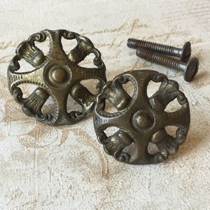 Peut inclure: Deux boutons de cabinet en laiton antique avec un design décoratif et orné. Les boutons sont circulaires avec un motif en forme de croix et quatre accents décoratifs autour du centre. Les boutons sont accompagnés de deux vis.