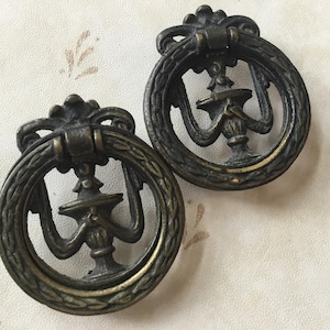 May include: Two antique brass drawer pulls with ornate designs. Each pull features a circular ring with a decorative floral pattern and a central design of a stylized urn with a flame.