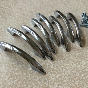 May include: A set of eight polished chrome cabinet handles with a curved design, arranged on a patterned surface. Small silver screws are visible to the right, suggesting a hardware set for furniture or cabinetry.