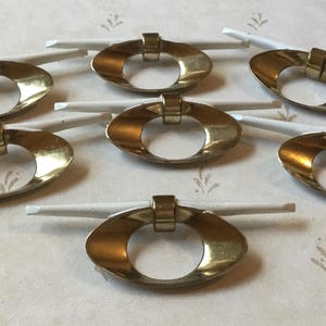 May include: Seven gold-toned oval drawer pulls with white rods, arranged in a circular pattern. The pulls have a shiny finish and a simple, elegant design. The background is a light-colored surface with a subtle floral pattern.