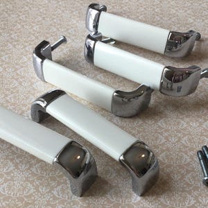 May include: Five cabinet handles with a white, rectangular central section and chrome-plated ends. They are arranged on a patterned surface, with a small group of screws. These are likely for kitchen or bathroom cabinets.