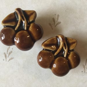 Op de afbeelding: Twee keramische kersvormige kastknoppen. Elke knop heeft drie bruine kersen met een glanzende afwerking en een donkerbruine steel en bladeren. De knoppen zijn geplaatst tegen een lichte achtergrond met een subtiel bladerpatroon.