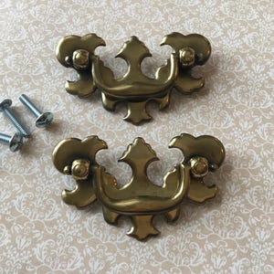 May include: Two antique brass drawer pulls with ornate designs, featuring a central decorative element and rounded ends. Four silver screws are included. The pulls are likely for furniture restoration or decorative projects.