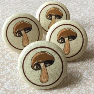 May include: Four round cabinet knobs with a mushroom design. Each knob has a cream background with a brown mushroom illustration inside a dark brown circular border. These decorative knobs are suitable for furniture.