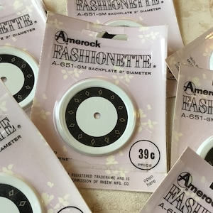 Peut inclure: Gros plan sur cinq plaques arrière en plastique blanc avec un motif en diamant noir et or. Les plaques arrière sont étiquetées "Amerock Fashionette A-651-GM Backplate 2" de diamètre. Une plaque arrière porte une étiquette de prix de 39 centimes.