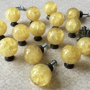 Peut inclure: Ensemble de boutons de meubles décoratifs avec un design de globe en verre craquelé jaune. Chaque bouton est monté sur une base en bronze foncé avec une vis argentée. Les boutons sont disposés sur une surface à motifs.