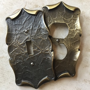 May include: Two antique brass wall plates, one with a single light switch and the other with a double outlet. The plates have a textured, hammered finish and ornate edges.
