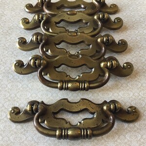 May include: A set of antique brass drawer handles. Each handle has an ornate design with a central grip and scrolled ends. The handles have a warm, aged patina, suggesting a vintage or retro style. They are made of brass.