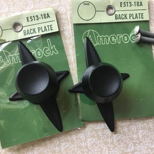 May include: Two matte black star-shaped cabinet knobs with four pointed arms, each mounted on a green cardboard backing card labeled Amerock E513-18A Back Plate. Two silver-toned metal screws are included with the right-hand knob for installation.