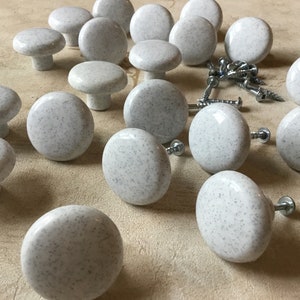 May include: A collection of 18 white ceramic cabinet knobs with a speckled grey finish. The knobs have a round shape and a small, raised centre. They are arranged on a light brown surface.