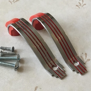 May include: Two vintage red and chrome cabinet door pulls with three red stripes on each. The pulls are curved and have a rounded edge. The pulls are on a white surface.
