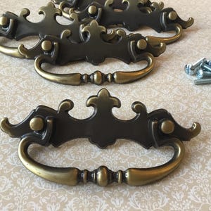 Puede incluir: Tiradores de cajón de estilo antiguo con acabado bronce oscuro y asas de latón. Los tiradores tienen un diseño ornamentado con curvas decorativas y un adorno central. Tornillos de montaje incluidos.