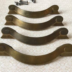 May include: Four antique brass drawer pulls with a curved design and decorative scroll ends. The pulls are in a warm, aged brass finish, with several screws visible at the top. These vintage hardware pieces are ideal for furniture restoration projects.