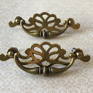 May include: Two antique brass drawer pulls with an ornate, symmetrical design. Each pull features a central floral-like motif and curved handles. The hardware is a warm, aged brass color, suggesting vintage or retro style.