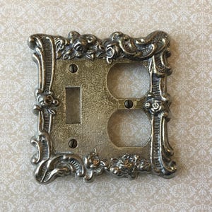 May include: A decorative silver-toned light switch cover with ornate floral and scrollwork detailing. The cover features a single toggle switch opening and two electrical outlet openings. The design is symmetrical and vintage-inspired.